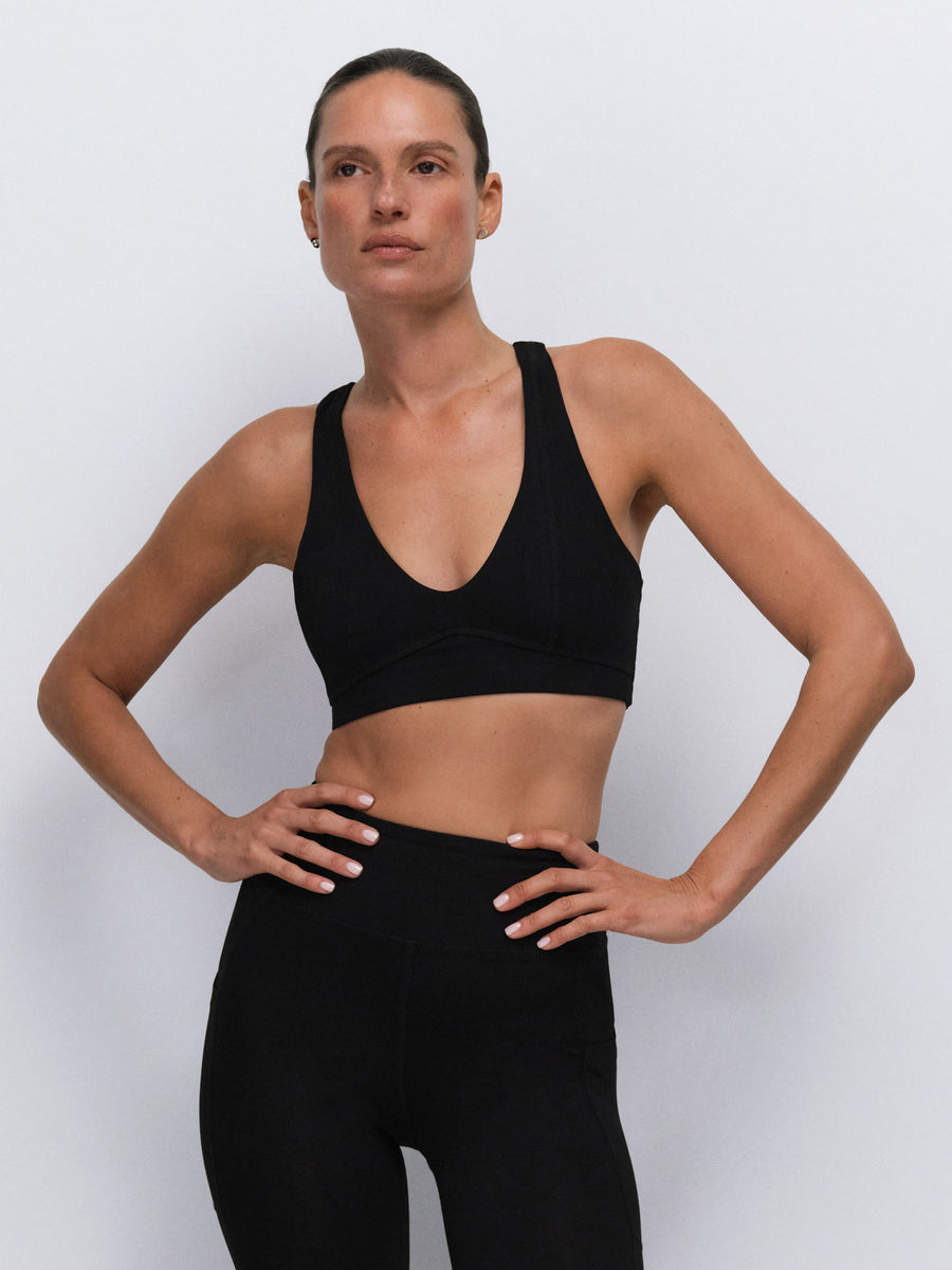 A woman modeling a black scoop-neck sports bra and matching black high-waisted leggings against a light gray studio background.