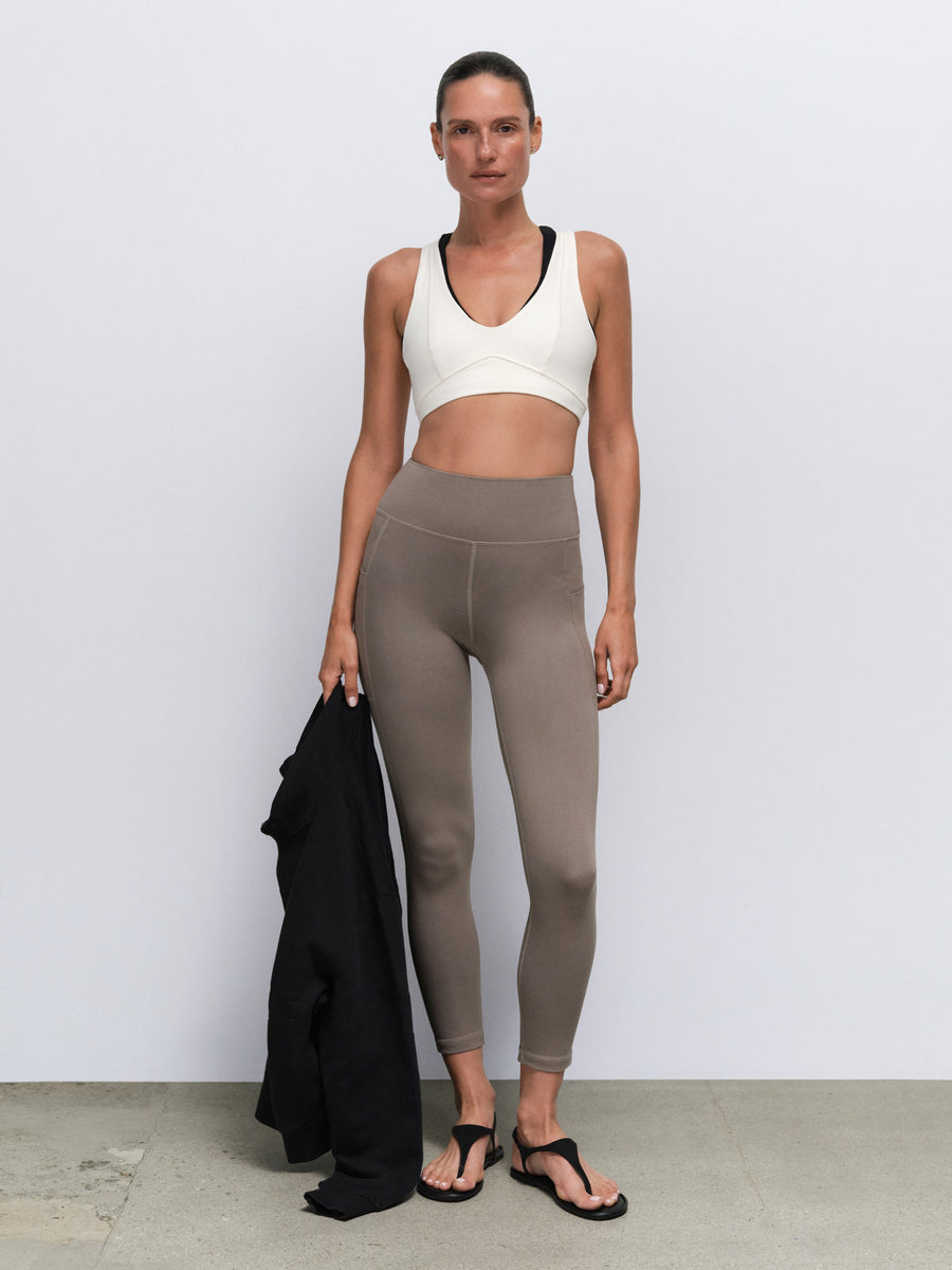 A woman stands against a light gray wall wearing a white sports bra with black trim and taupe high-waisted leggings, holding a black cardigan. She is wearing black flip-flop sandals.