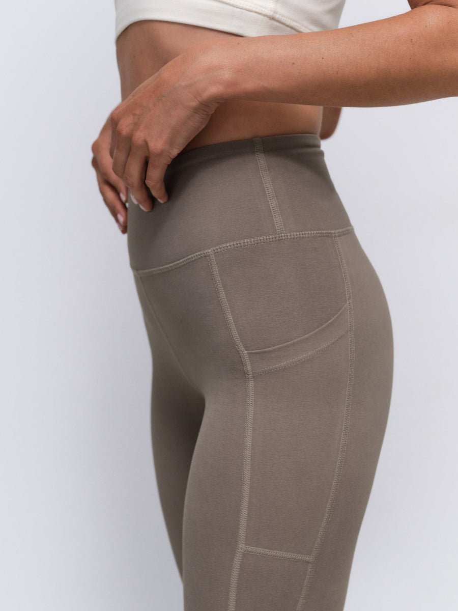 Close-up side view of a person wearing high-waisted taupe leggings with seam detailing and a side pocket, paired with a white cropped top against a light gray studio background.