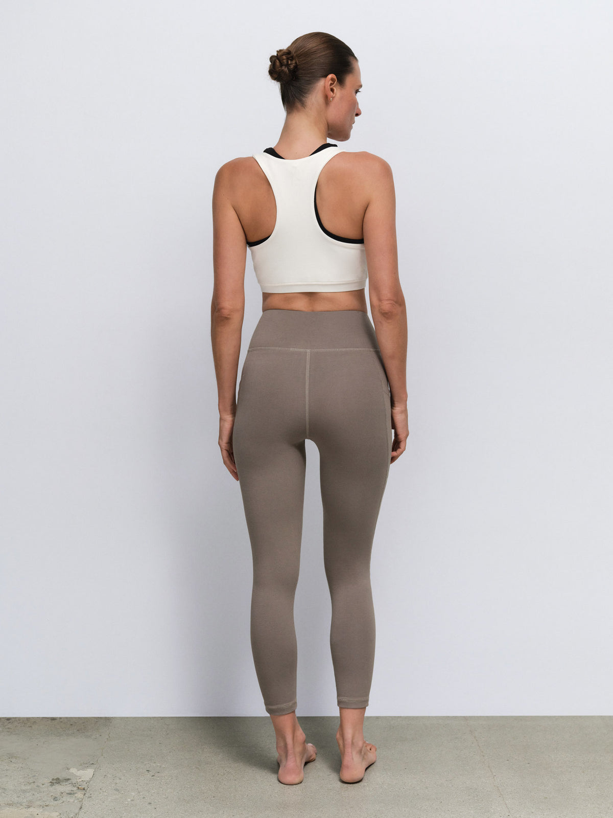 A woman viewed from behind wearing a white sports bra with black trim and high-waisted taupe leggings, standing against a plain light gray wall.