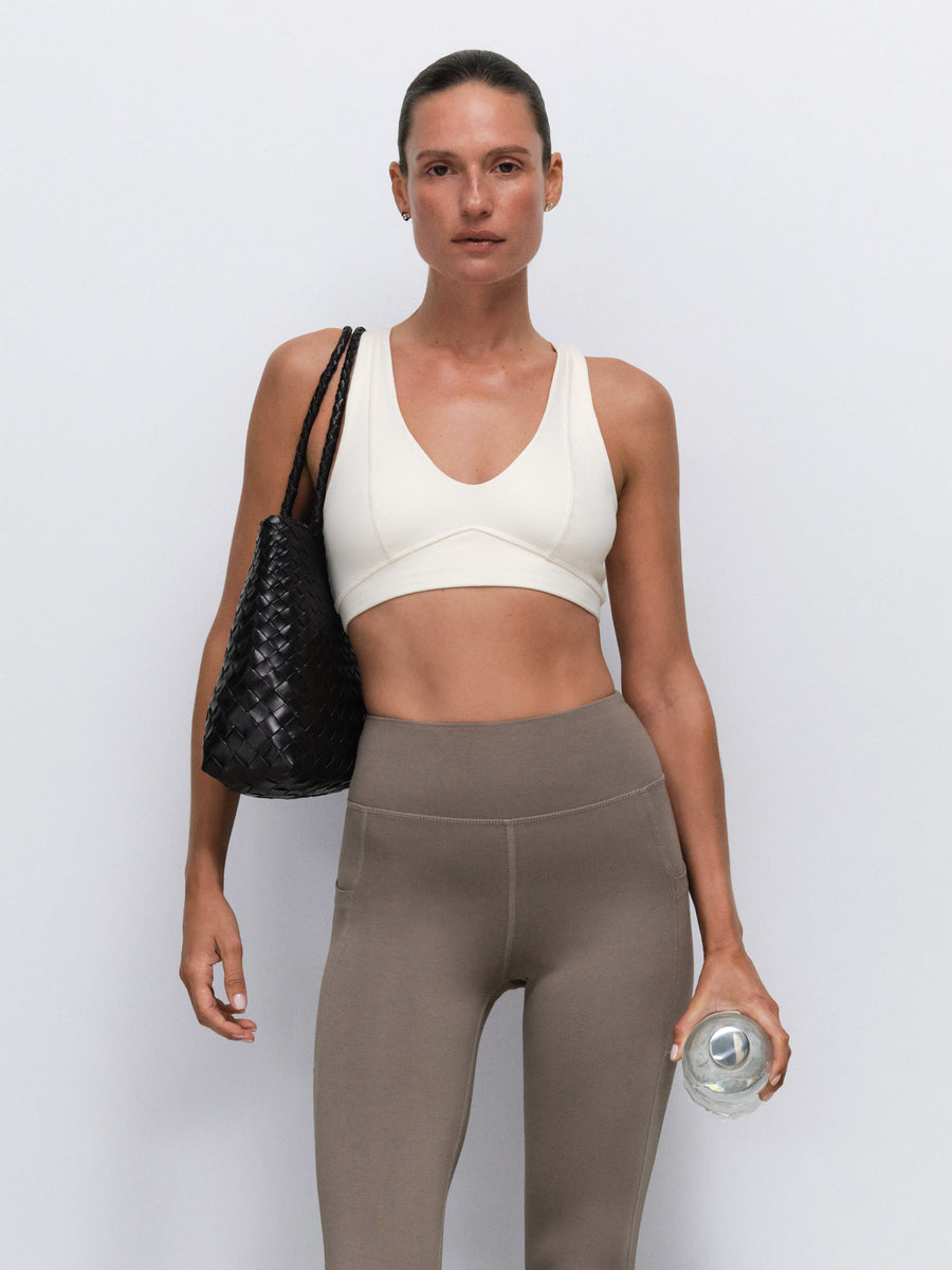 A woman modeling a white sports bra and taupe high-waisted leggings, carrying a black woven shoulder bag and holding a clear water bottle against a light gray studio background.