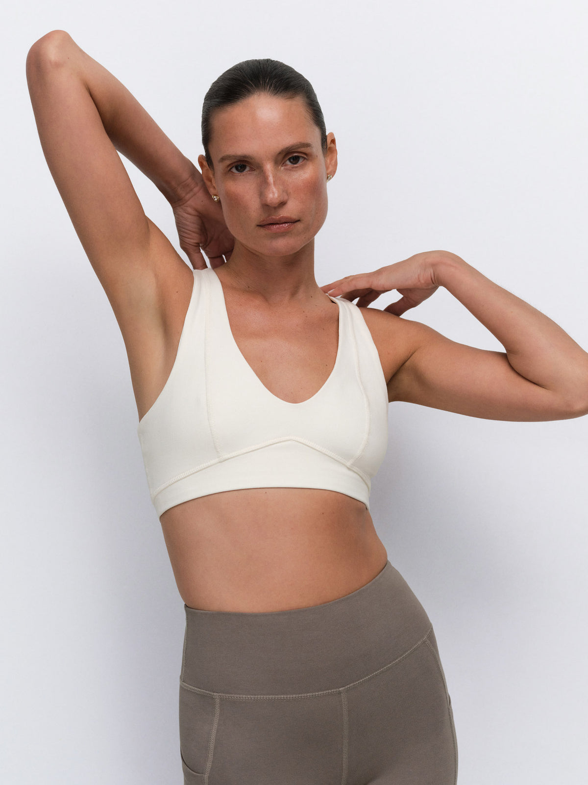 A woman modeling a cream-colored sports bra and taupe-gray high-waisted leggings against a light gray studio background.