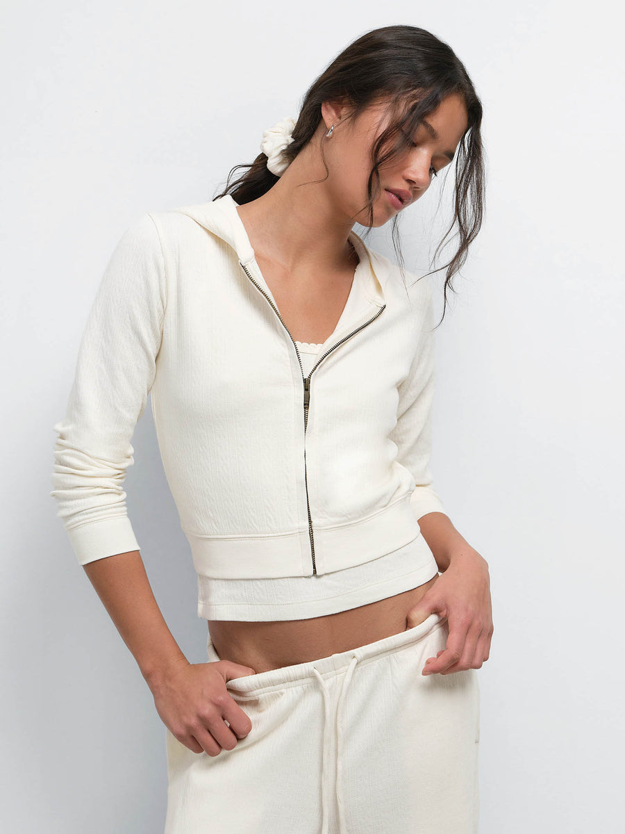 Model looking down and adjusting the waistband of the Organic Pointelle Pant in Cream, wearing the matching Shrunken Hoodie