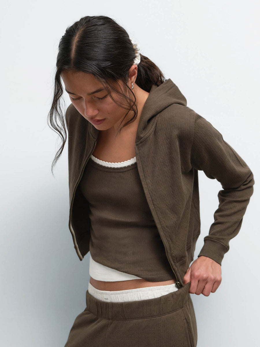 Model looking down while zipping the Organic Pointelle Shrunken Hoodie in Mocha over a matching tank and jogger