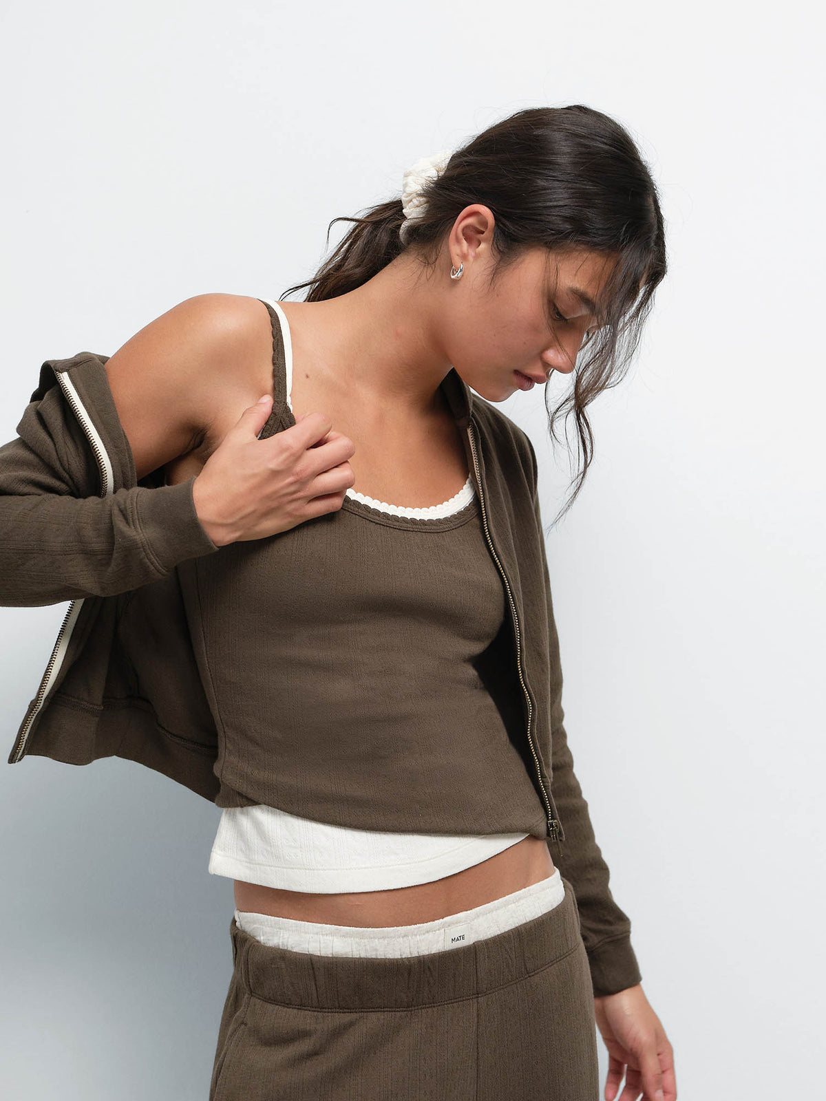 Model slipping off the Organic Pointelle Shrunken Hoodie in Mocha to reveal a cream-trimmed tank and matching jogger