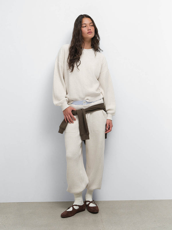 Full-length view of model with a brown sweater tied at the waist over the Organic Waffle Crewneck Sweatshirt and Sweatpant in Oat