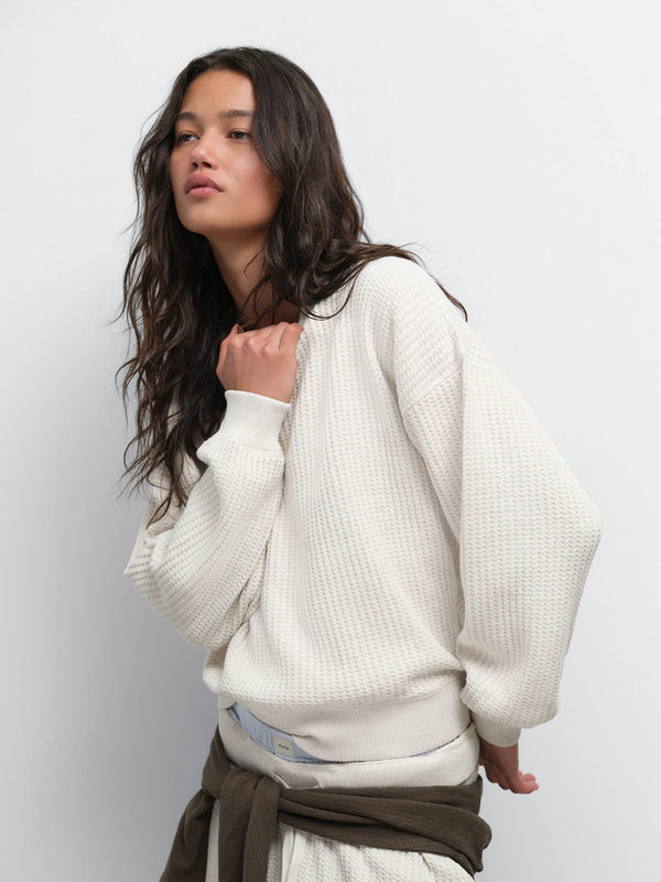 Model touching her collar, wearing the Organic Waffle Crewneck Sweatshirt in Oat with a brown sweater tied at the waist