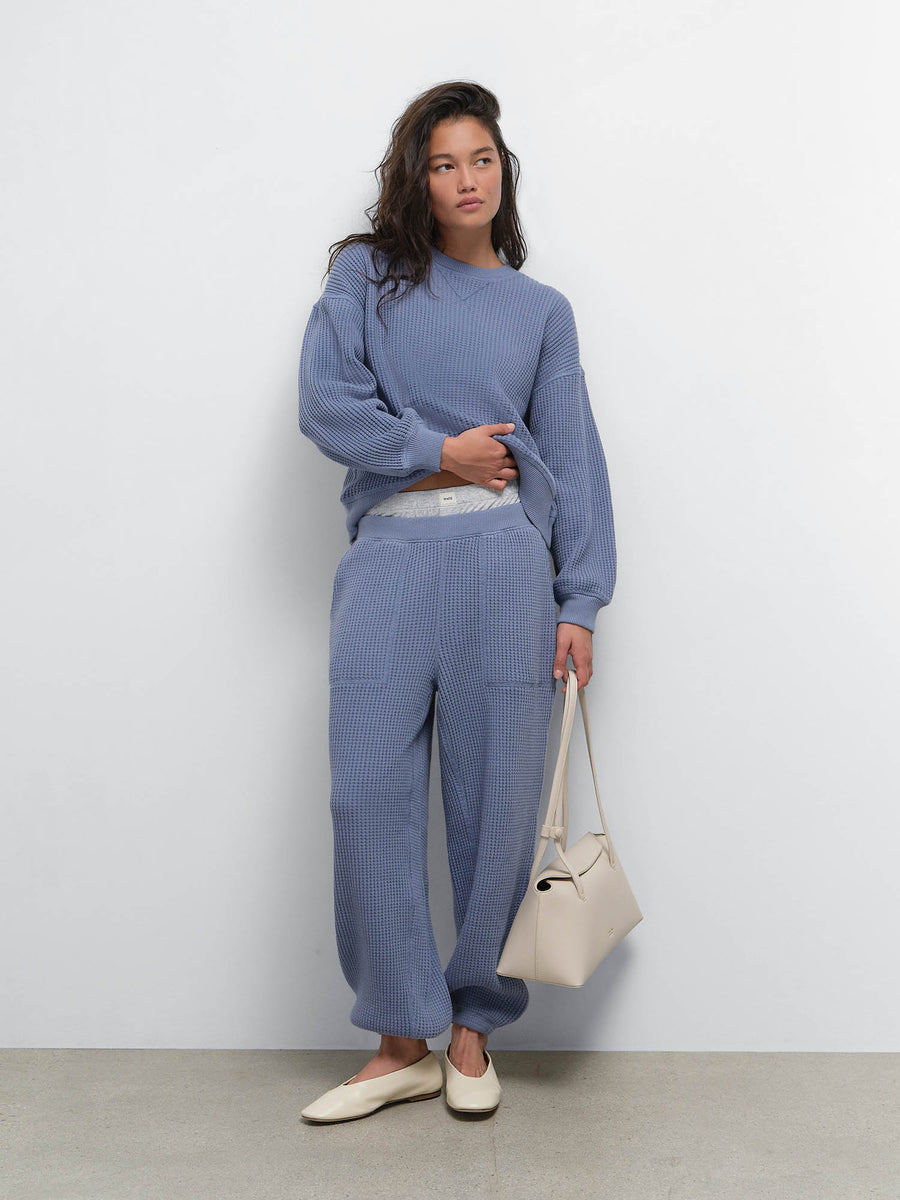 Full-length view of model holding a cream bag in the Organic Waffle Crewneck Sweatshirt and Sweatpant in Bay