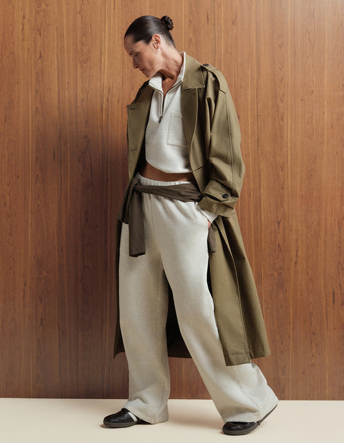 MATE model wearing an olive green trench coat over a heather oat wide-leg pant set, standing against a wood-paneled wall