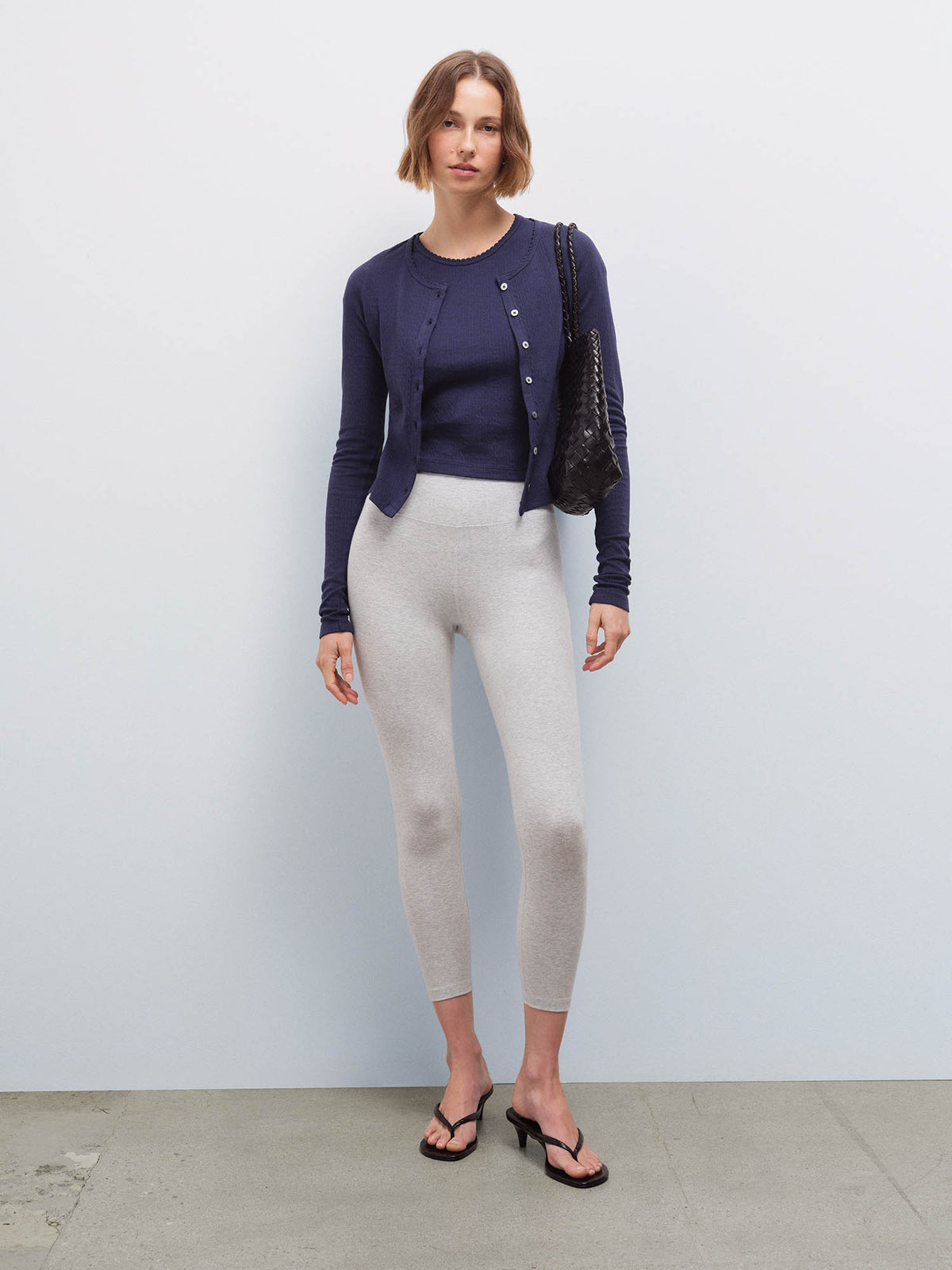 Full-length front view of a woman wearing a navy pointelle cardigan and heather grey leggings, carrying a black bag