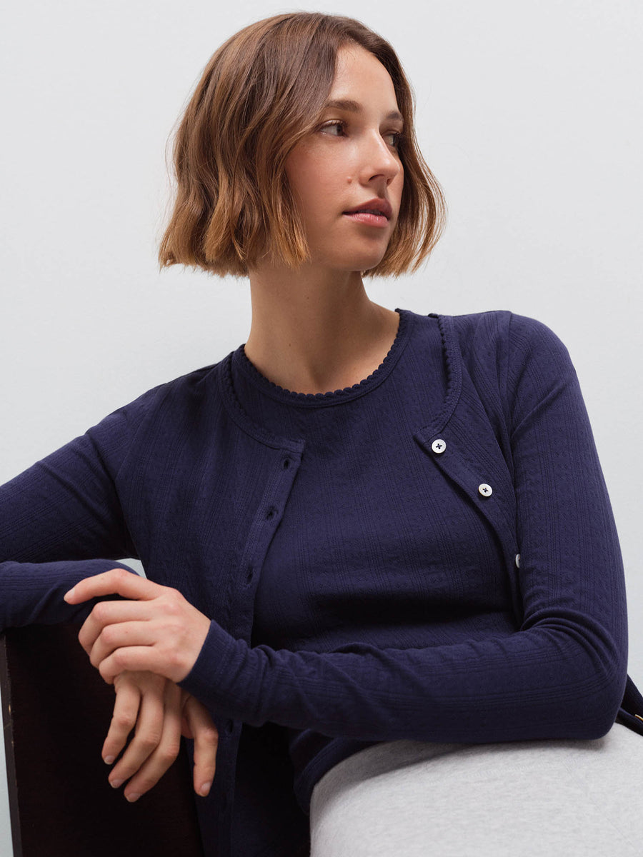 Woman seated leaning on a chair wearing a navy pointelle cardigan over a matching tank top