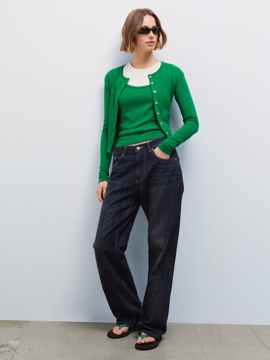 Full-length front view of a woman wearing a green pointelle cardigan over a cream tank with dark denim jeans and sunglasses