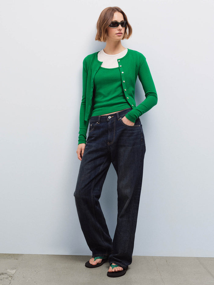 Full-length front view of a woman wearing a green pointelle cardigan over a cream tank with dark denim jeans and sunglasses