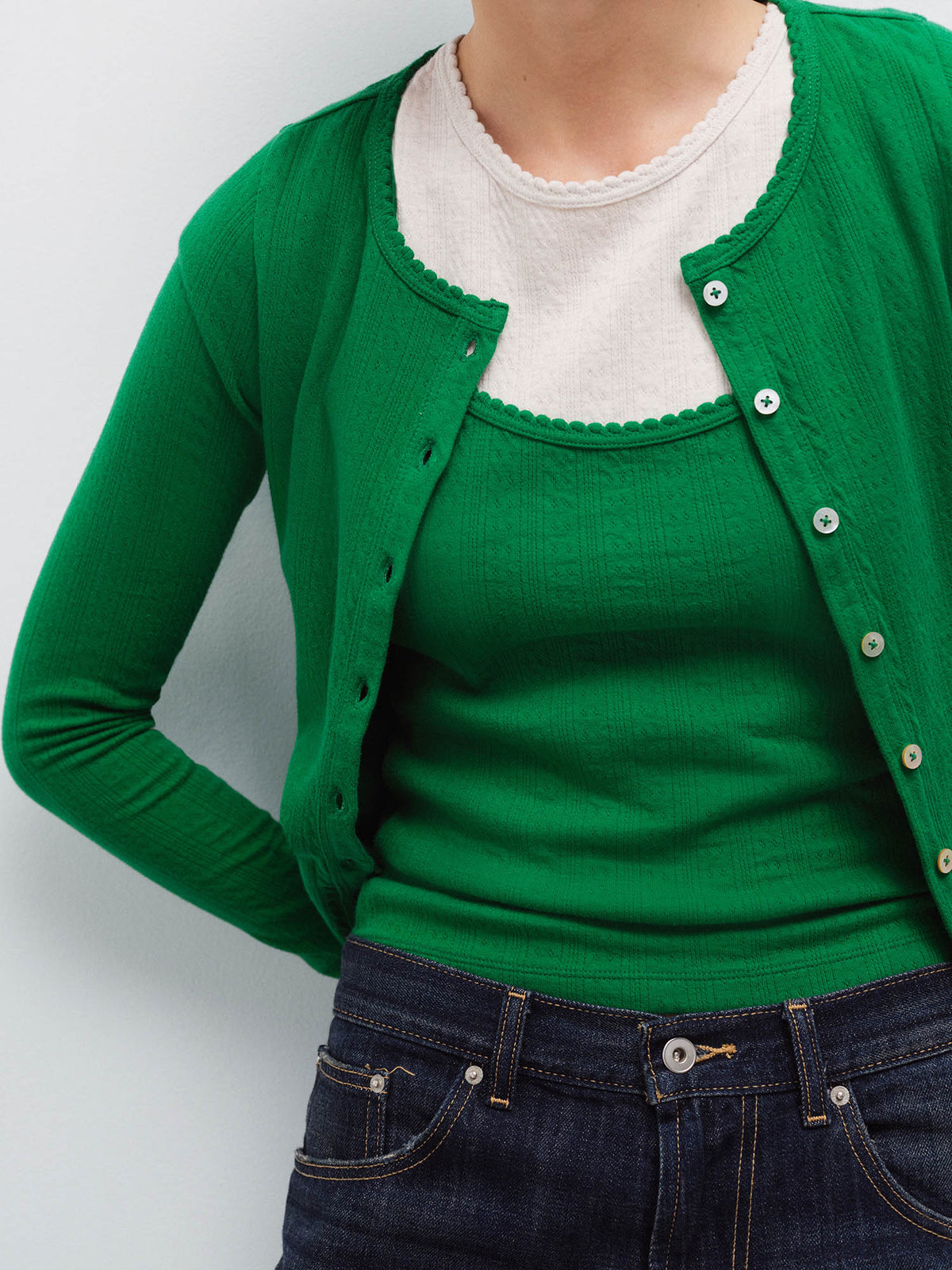 Close-up of a woman wearing an open green pointelle cardigan over a cream scalloped-trim tank with denim jeans