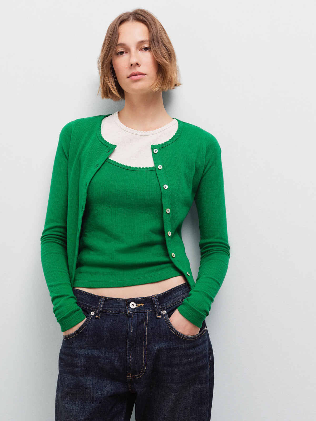 Woman wearing an open green pointelle cardigan over a cream tank with denim jeans, hands in pockets