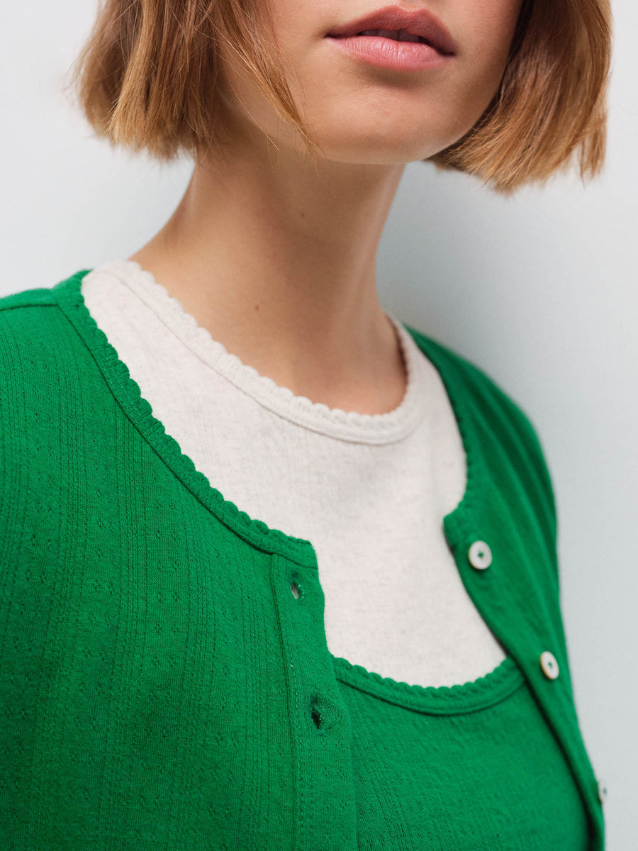 Close-up of a woman's neckline wearing a green pointelle cardigan over a cream scalloped-trim tank