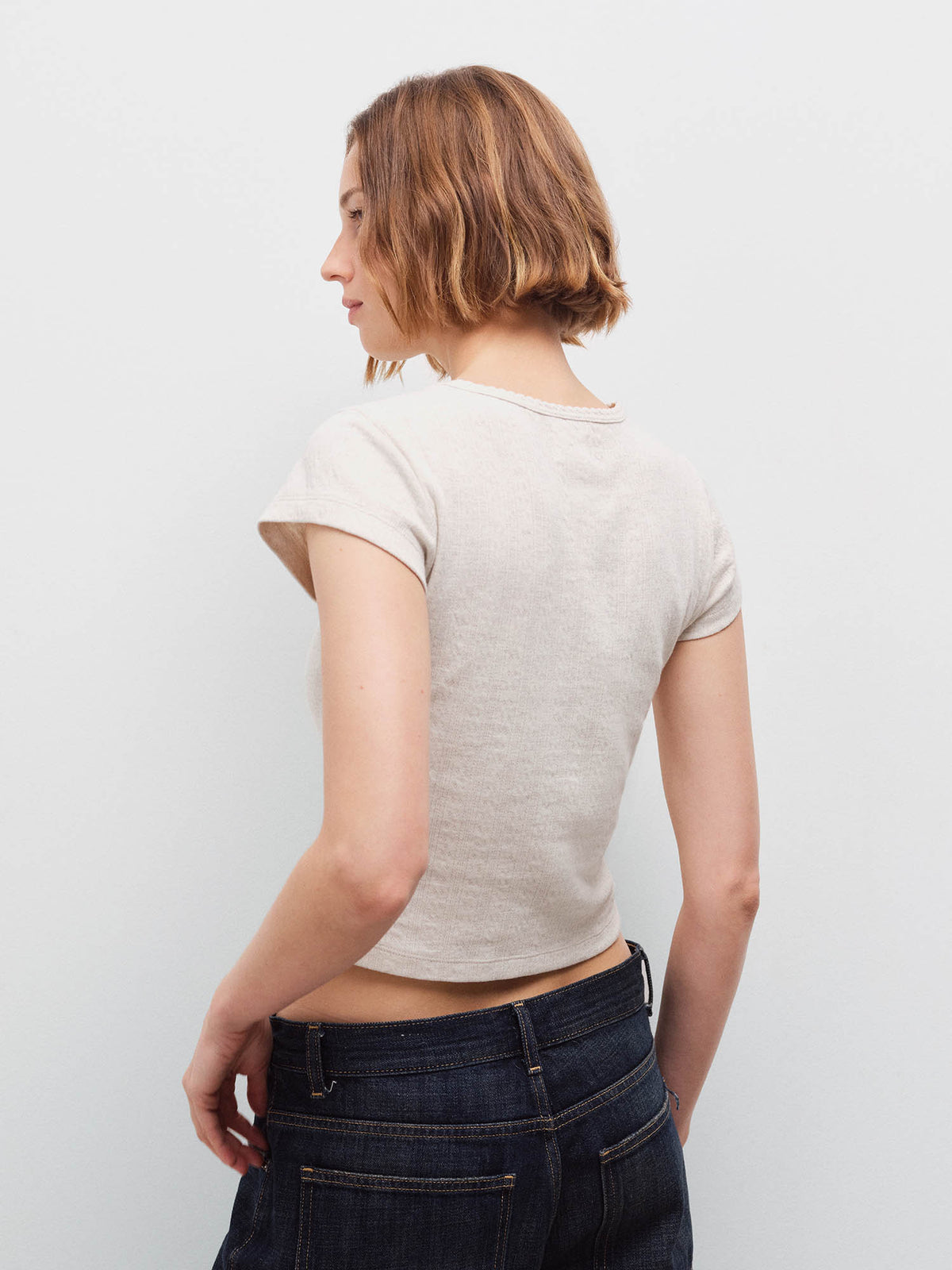 Woman standing wearing a cream cap-sleeve baby tee, back view with dark denim jeans