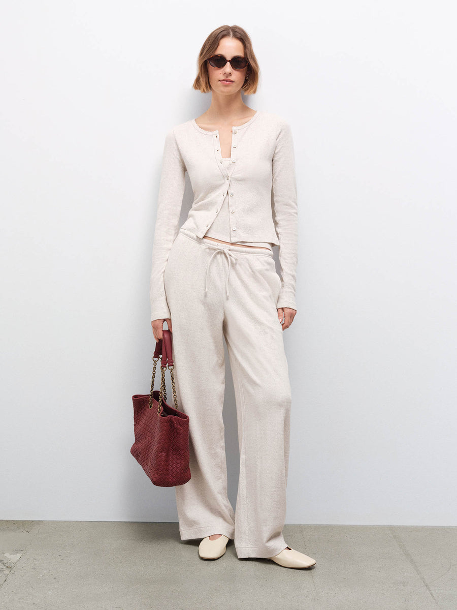 Woman wearing cream pointelle cardigan and matching wide-leg pants, holding a burgundy bag, full-length in studio