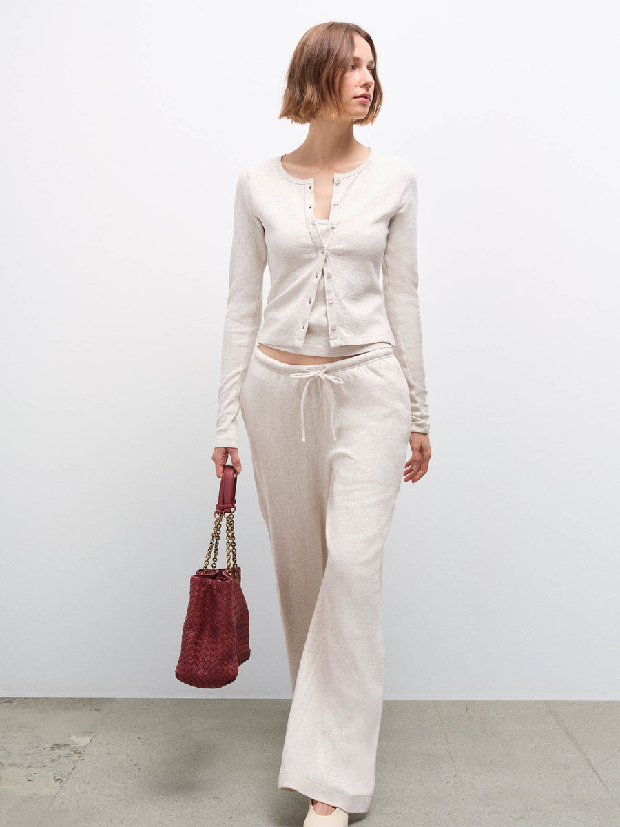 Woman wearing cream pointelle cardigan and wide-leg pants, walking while holding a burgundy bag in studio