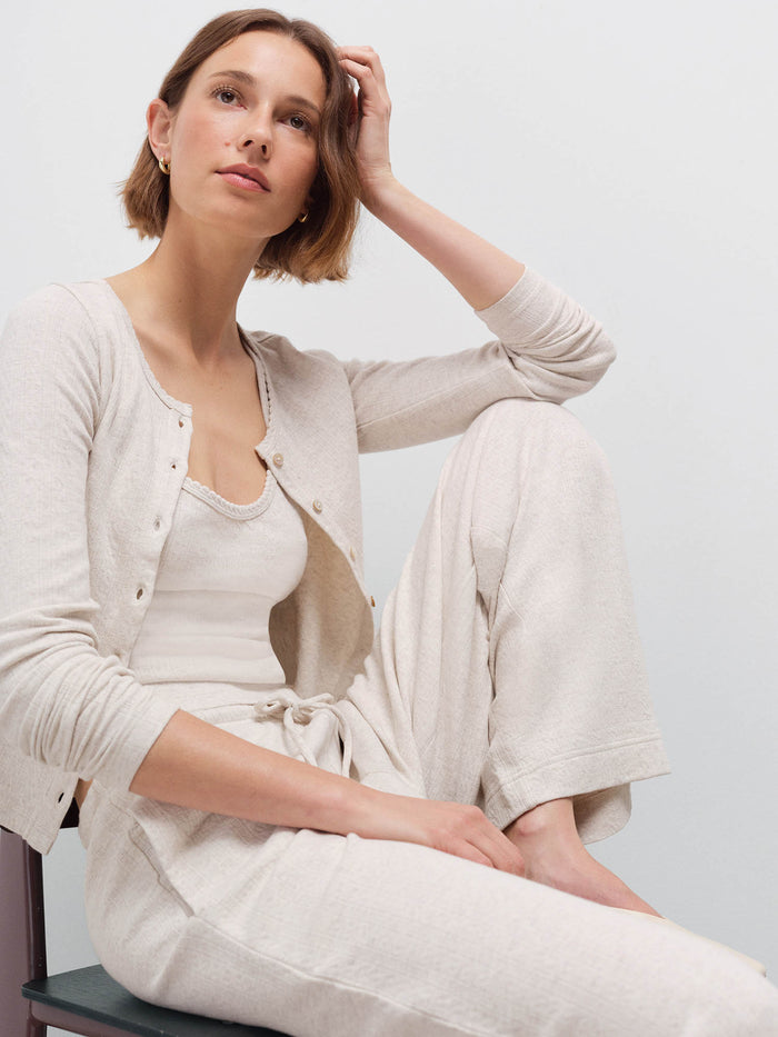 Woman wearing cream pointelle cardigan over matching tank and pants, seated with hand in hair in studio