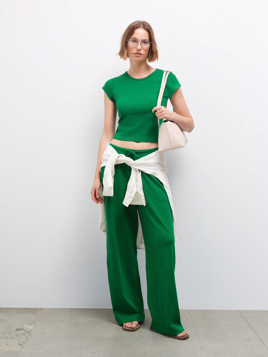 Woman wearing a green cropped top and wide-leg pants with a cream cardigan tied at the waist, carrying a cream bag