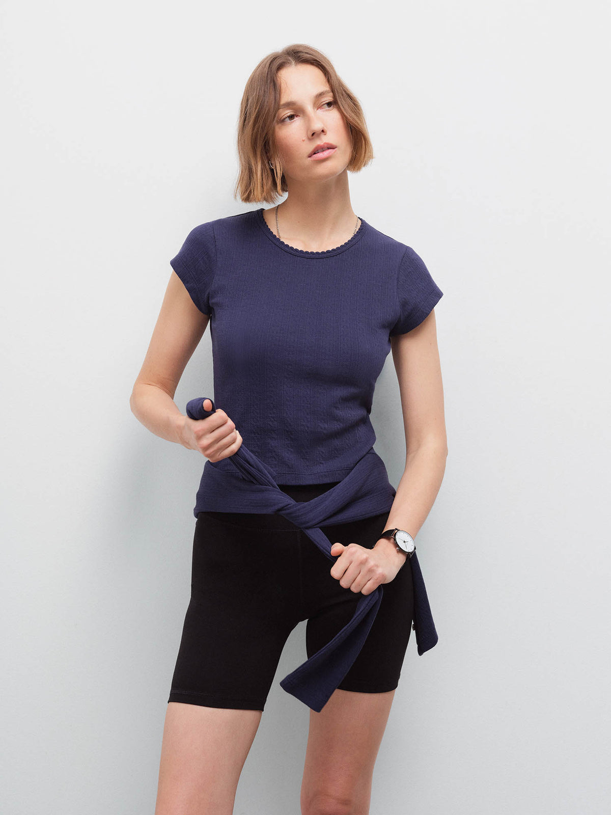 Woman wearing a navy pointelle top tied at the waist over black biker shorts, holding the tie ends