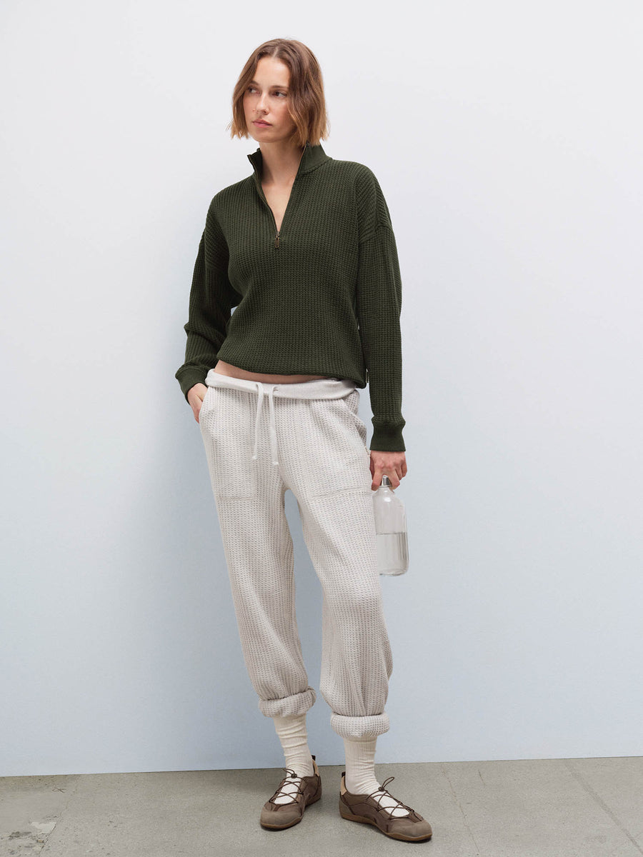 Woman wearing cream waffle-knit sweatpants and olive half-zip sweater, front view holding a water bottle in studio