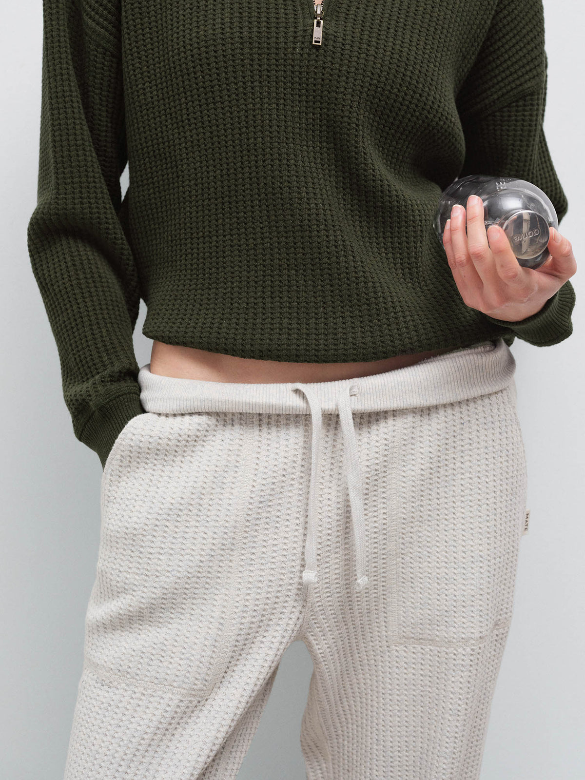 Close-up of cream waffle-knit sweatpants with olive half-zip sweater hem, holding a small object in studio