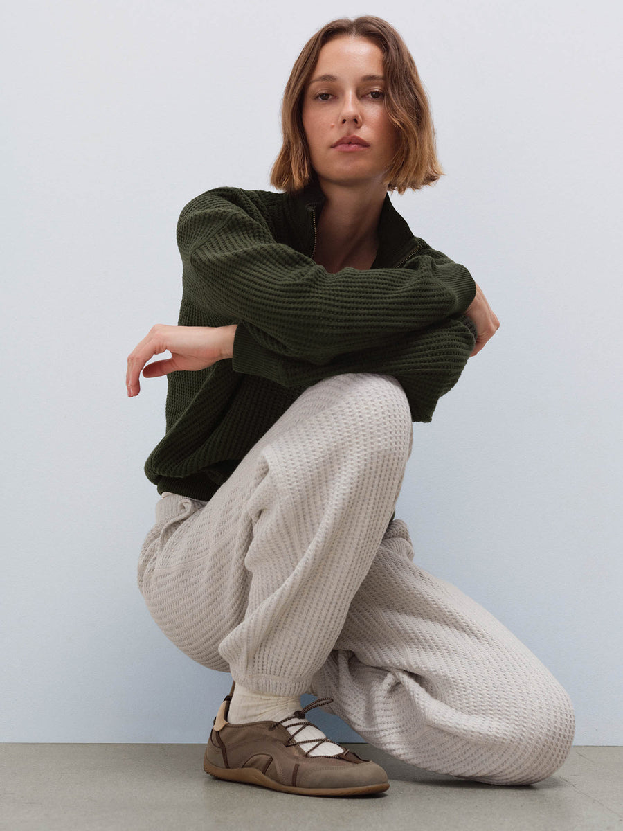 Woman wearing cream waffle-knit sweatpants and olive half-zip sweater, crouching pose in studio