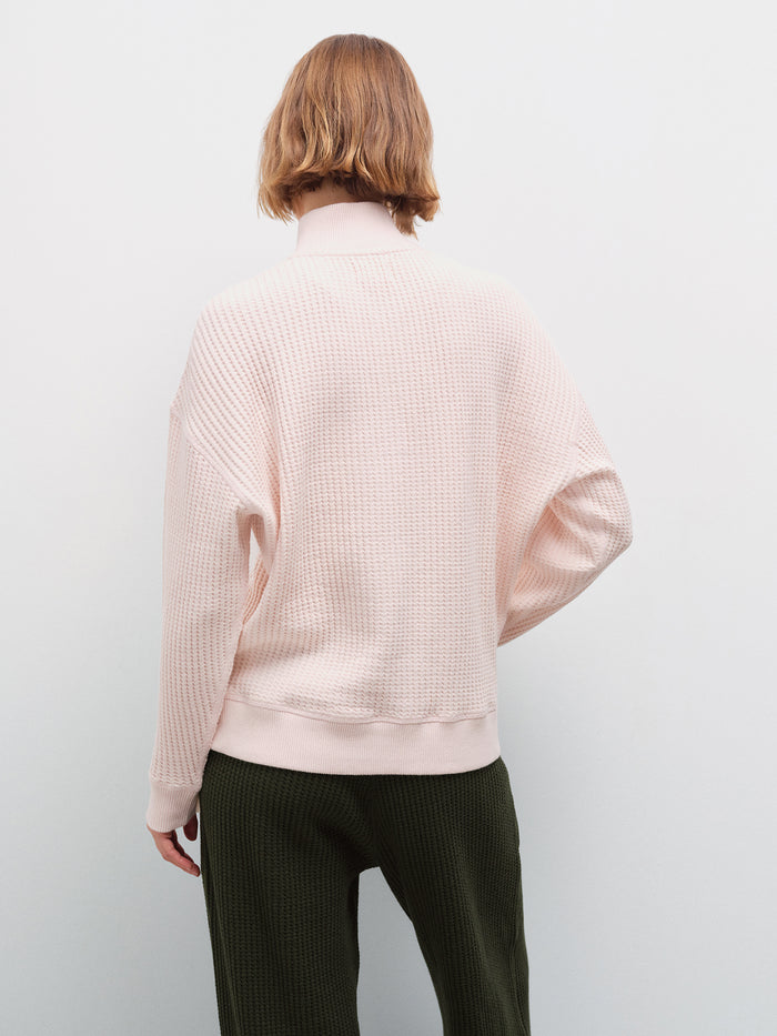 Woman standing wearing an oversized pale pink waffle-knit quarter-zip pullover and olive green pants, back view