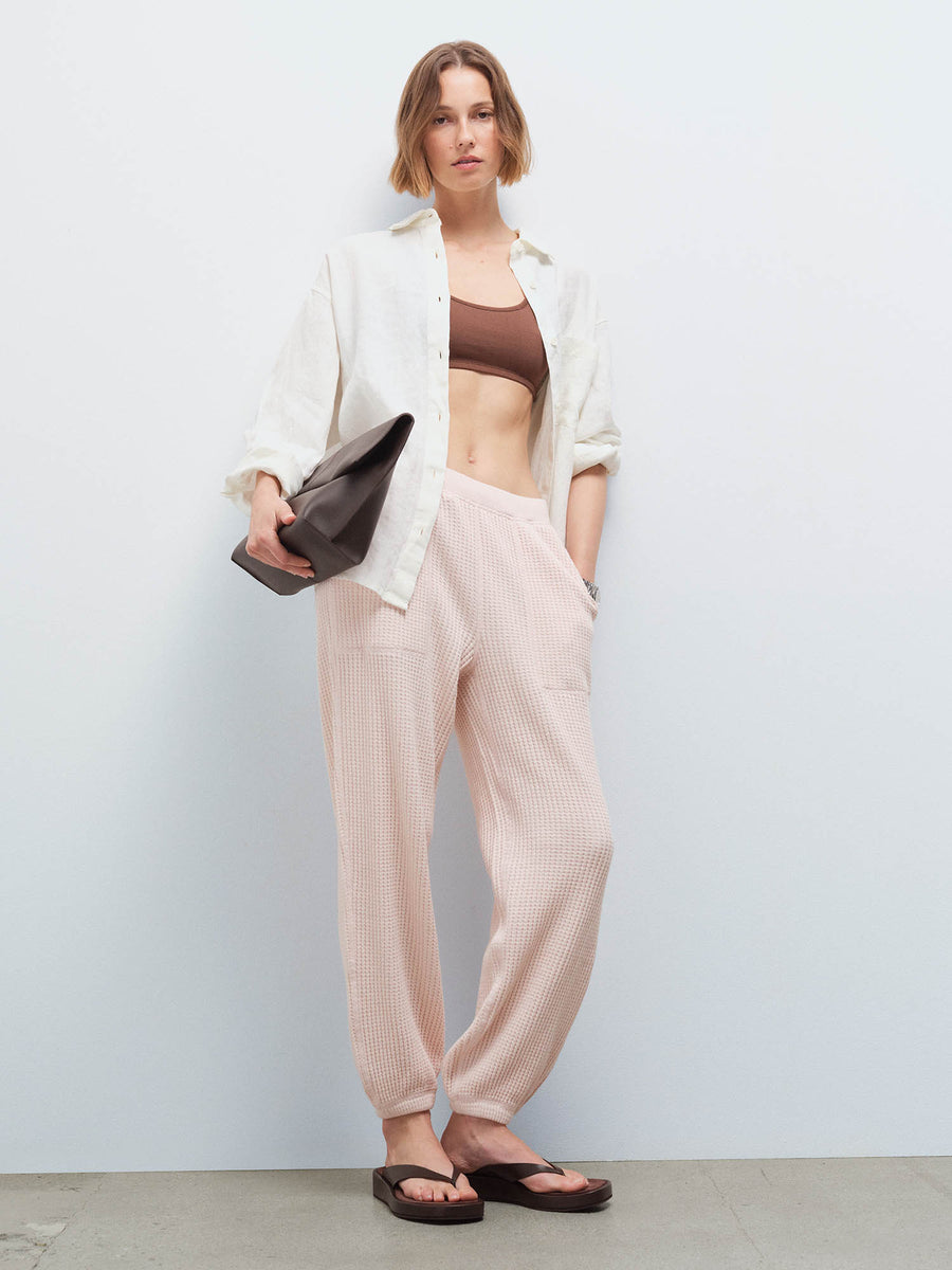 Woman wearing a cream open shirt over a brown bralette and pale pink waffle jogger sweatpants, carrying a dark clutch