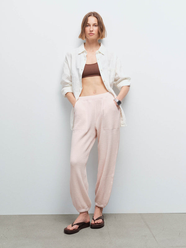 Woman wearing an open cream shirt over a brown bralette and pale pink waffle jogger sweatpants with sandals, studio