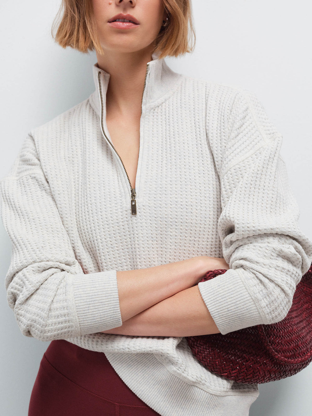Close-up of a woman with arms crossed wearing a cream waffle-knit quarter-zip pullover, holding a woven bag