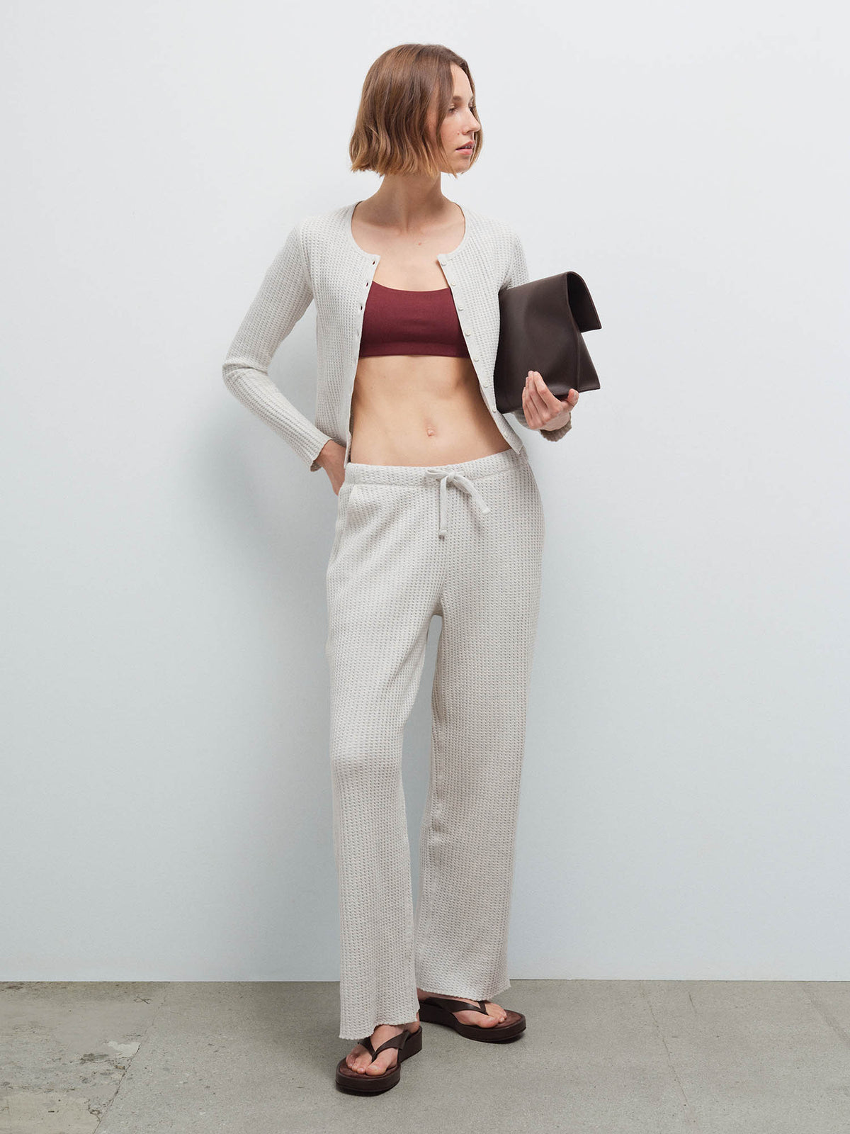 Woman wearing an open cream waffle-knit cardigan over a burgundy bralette with matching pants, holding a dark clutch