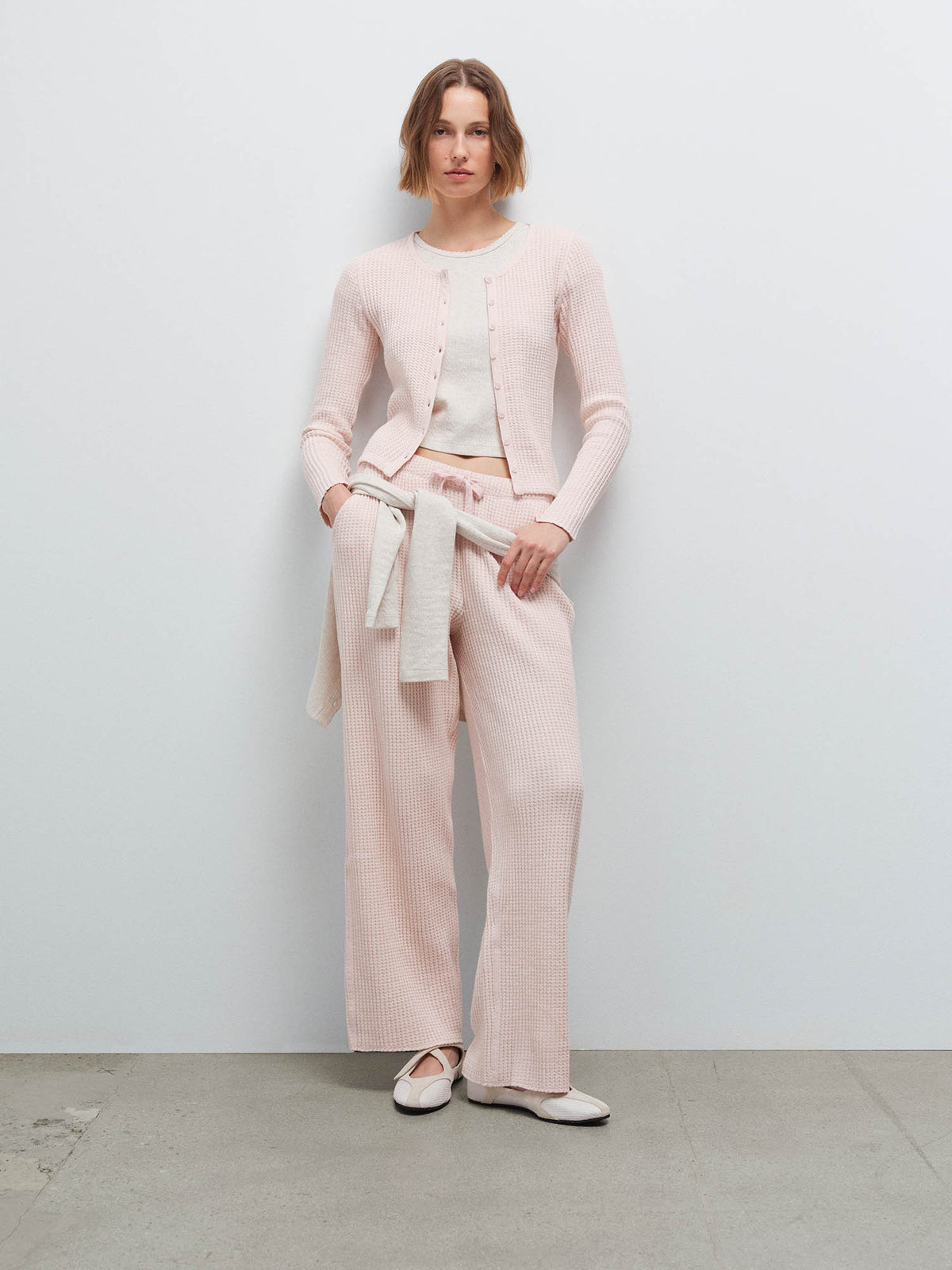 Woman wearing a pale pink waffle-knit cardigan and pants with a cream sweater tied at the waist, hand in pocket
