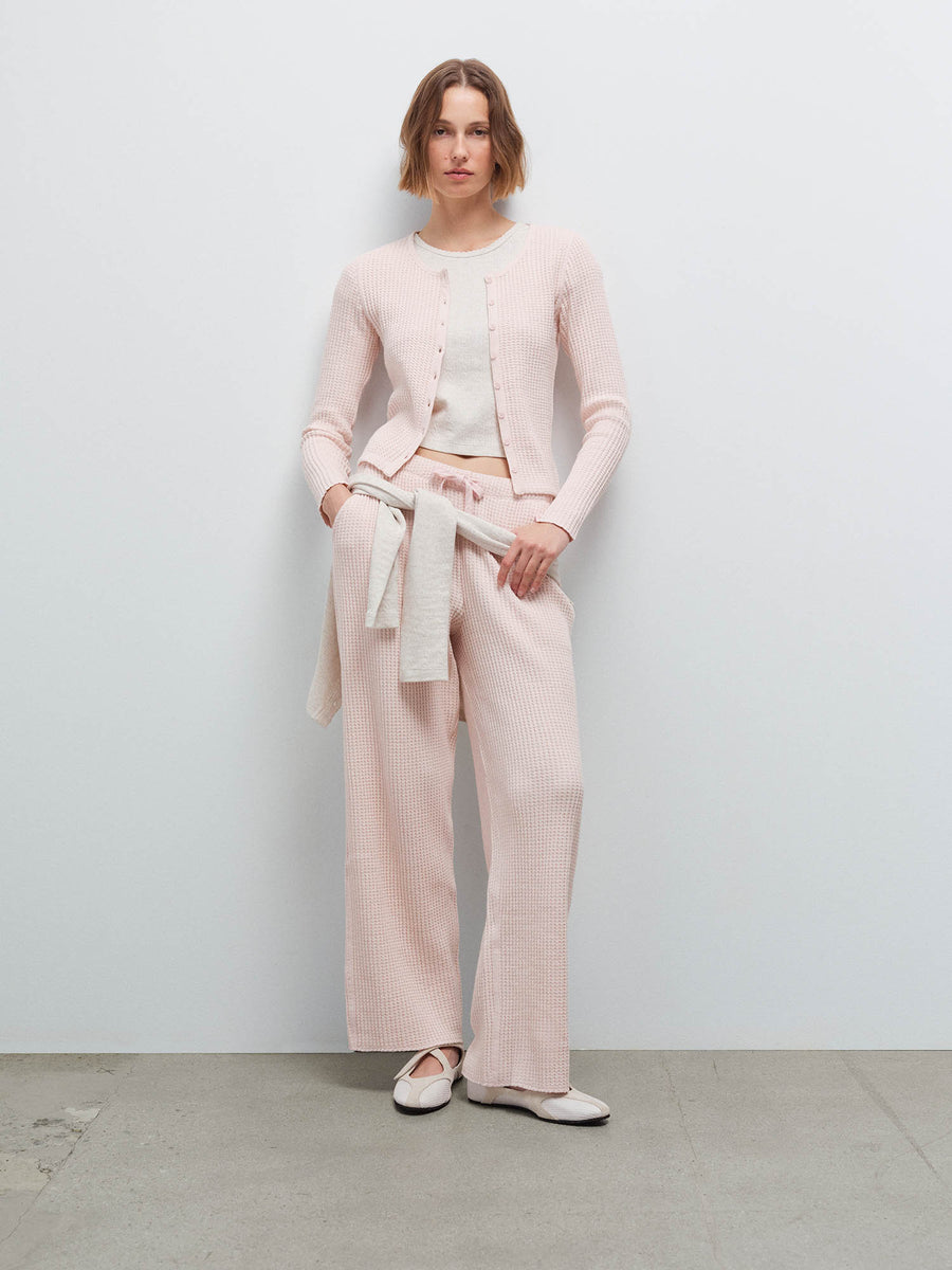 Woman wearing a pale pink waffle-knit cardigan and pants with a cream sweater tied at the waist, hand in pocket