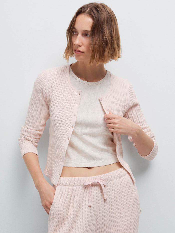 Woman wearing a pale pink waffle-knit cropped cardigan over a cream tank with matching joggers, studio
