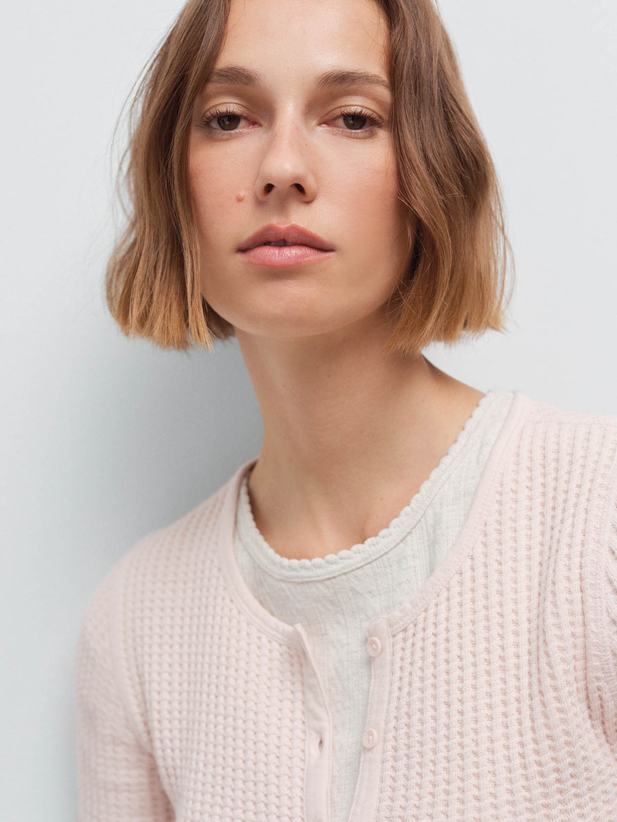 Close-up of a woman's face wearing a pale pink waffle-knit cardigan over a cream scalloped-trim top, studio