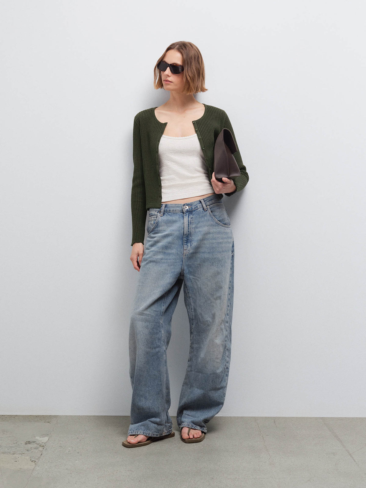 Woman wearing an open olive green waffle-knit cardigan over a cream top with wide-leg jeans and sunglasses, holding a clutch