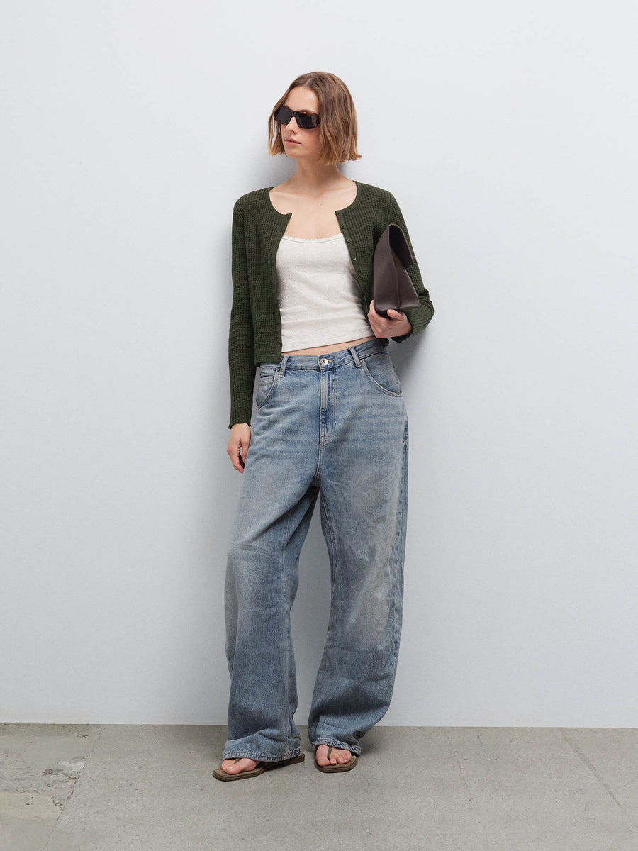 Woman wearing an open olive green waffle-knit cardigan over a cream top with wide-leg jeans and sunglasses, holding a clutch