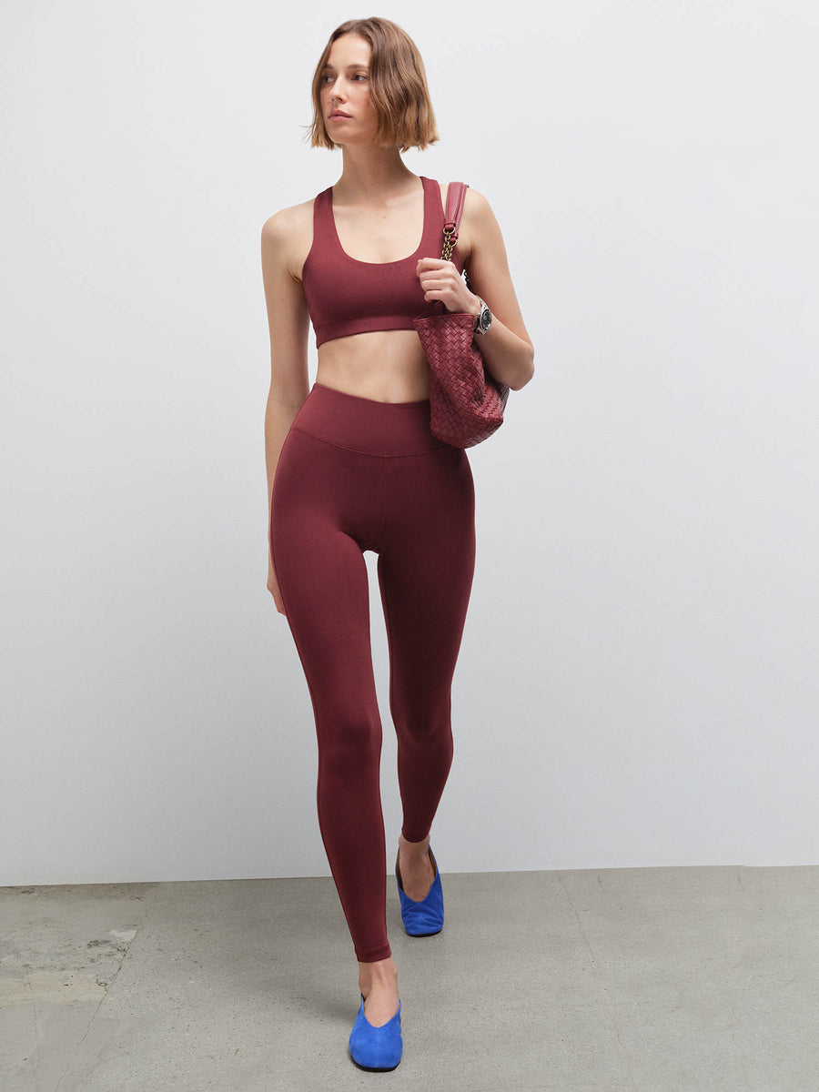 Woman walking wearing a burgundy racerback sports bra and matching leggings with blue flats, carrying a red bag