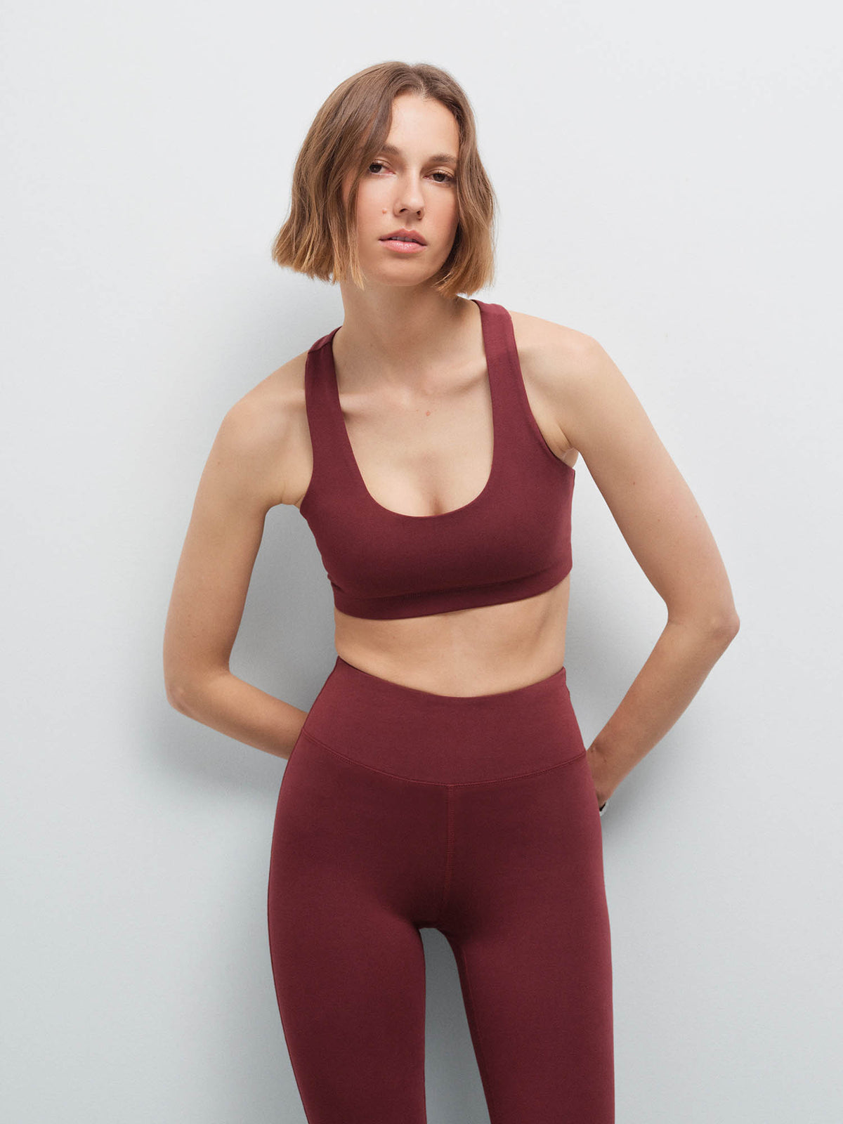 Woman standing with hands behind her back wearing a burgundy racerback sports bra and matching leggings