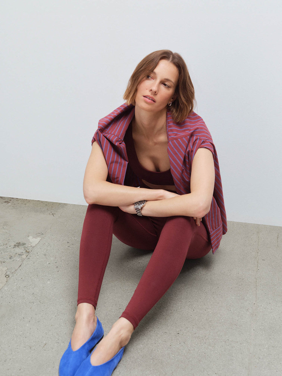 Woman seated wearing a red striped shirt over a burgundy sports bra and matching leggings with blue flats