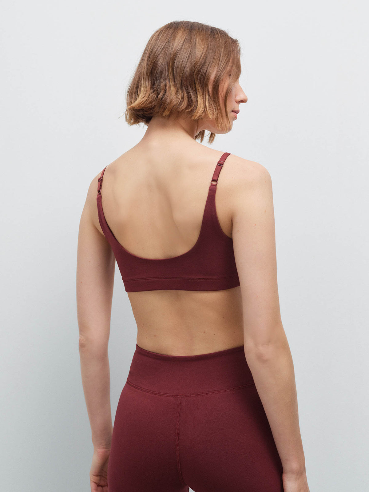 Woman standing wearing a burgundy scoop-neck bra and matching leggings, back view in studio