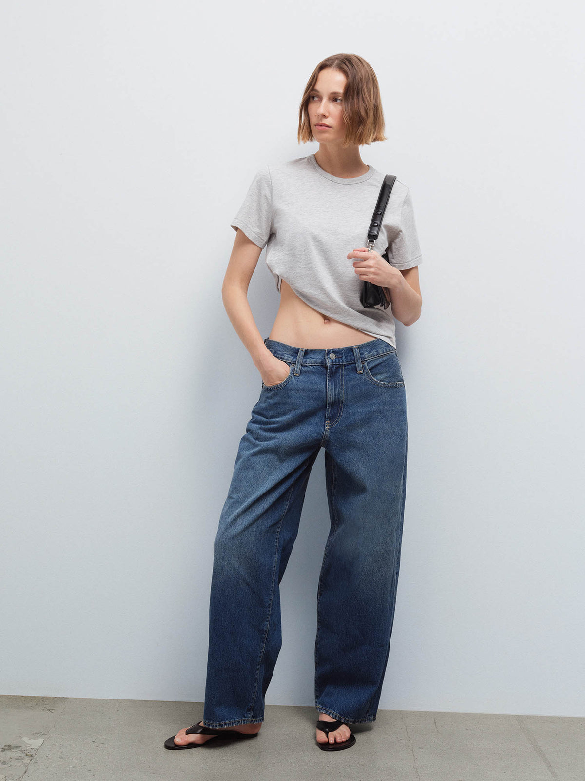 Full-length front view of a woman wearing a cropped heather grey tee and wide-leg denim jeans, holding a black bag