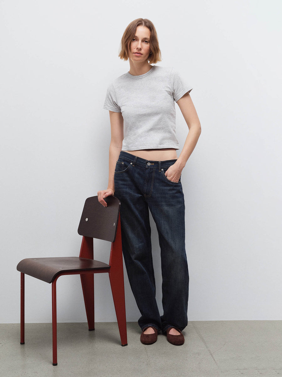 Full-body front view of a woman wearing a light heather gray cropped tee and wide-leg dark denim jeans beside a chair