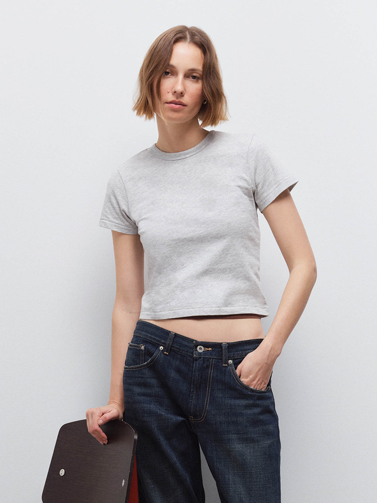 Woman wearing a light heather gray cropped tee and dark denim jeans, holding a dark bag, front view in studio