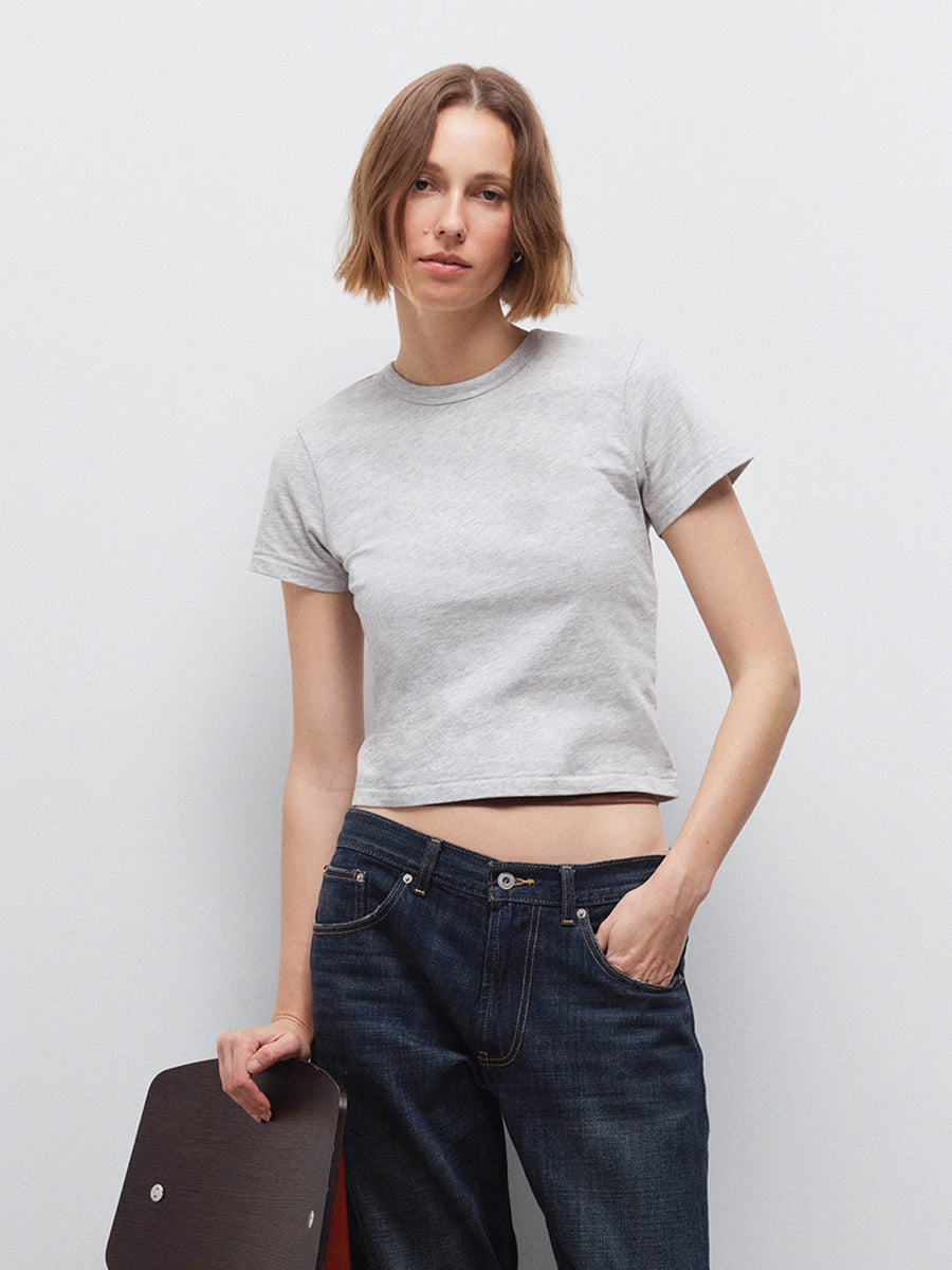 Woman wearing a light heather gray cropped tee and dark denim jeans, holding a dark bag, front view in studio