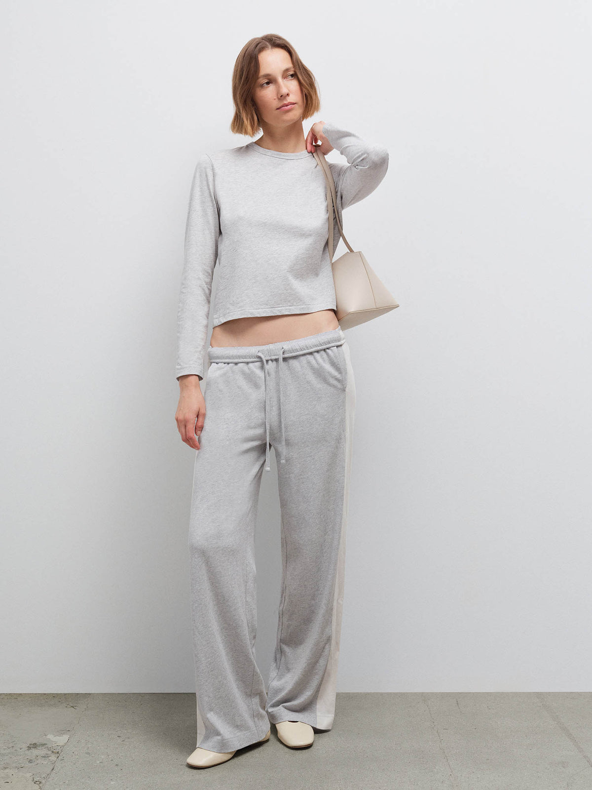 Full-length front view of a woman wearing a heather grey long-sleeve top and matching wide-leg sweatpants, holding a bag