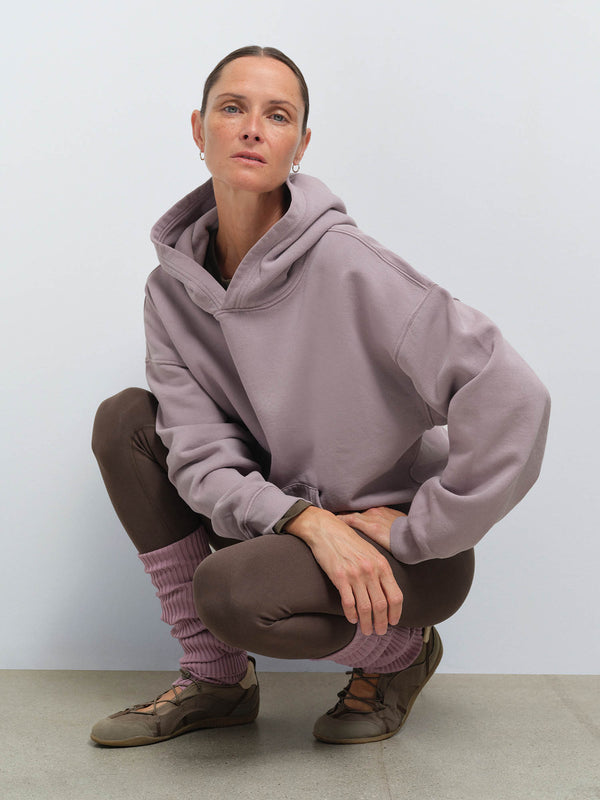 MATE model crouching wearing an oversized mauve organic fleece pocket hoodie, brown leggings, mauve leg warmers, and brown lace-up shoes against a light background